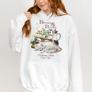 May include: White hooded sweatshirt with a graphic print of a cat sleeping on a stack of books, a glass bottle, and a sprig of green leaves. The text "Brews & Books" is above the image and "Enchanting Reads Cozy Days" is below the image.