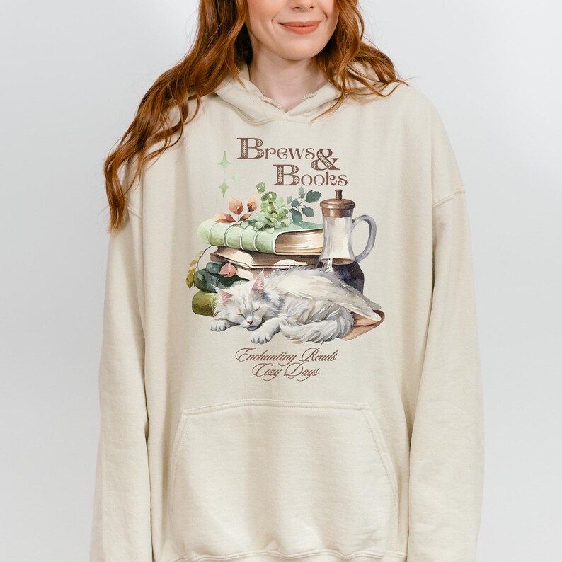 May include: A cream-colored hoodie with a graphic print of a cat sleeping on a stack of books. The graphic also includes a glass bottle and the text "Brews & Books" and "Enchanting Reads Cozy Days".