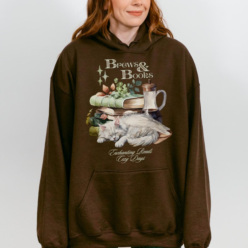 May include: A brown hoodie with a white cat sleeping on a stack of books. The hoodie has the text "Brews & Books" and "Enchanting Reads Cozy Days" printed on it.
