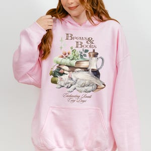 May include: A pink hoodie with a graphic of a white cat sleeping on a stack of books, a glass bottle, and green vines. The text "Brews & Books" is above the graphic, and the text "Enchanting Reads Cozy Days" is below the graphic.
