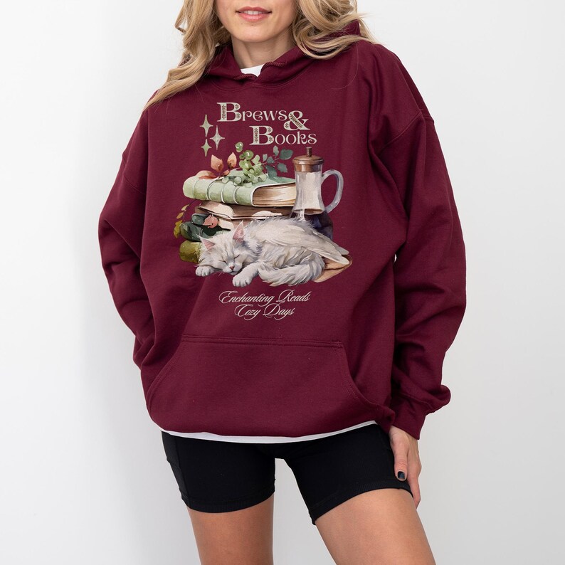 May include: A maroon hoodie with a white cat sleeping on a stack of books. The hoodie has the text "Brews & Books" and "Enchanting Reads Cozy Days" printed on it.