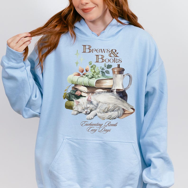 May include: Light blue hooded sweatshirt with a graphic design of a cat sleeping on a stack of books, a glass pitcher, and grapes. The text "Brews & Books" is above the graphic and "Enchanting Reads Cozy Days" is below the graphic.