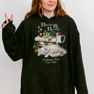 May include: Black hooded sweatshirt with a graphic print of a cat curled up with a stack of books and a mug. The text on the graphic reads "Brews & Books", "Enchanting Reads", and "Cozy Days".
