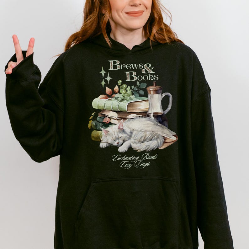 May include: Black hooded sweatshirt with a graphic print of a cat curled up with a stack of books and a mug. The text on the graphic reads "Brews & Books", "Enchanting Reads", and "Cozy Days".