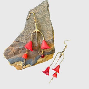 Puede incluir: Un par de pendientes colgantes en tono dorado con múltiples cuentas en forma de flor roja suspendidas de finas barras metálicas. Cada pendiente incluye un cierre de gancho, delicadas cadenas metálicas y pequeñas cuentas rojas facetadas.