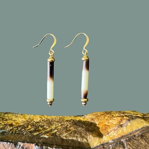 May include: A pair of handmade earrings featuring a unique design. Each earring has a gold-colored hook and a cylindrical pendant with alternating bands of dark brown and cream colors. The earrings are suspended from gold-colored accents.