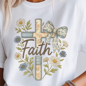 以下が含まれることがあります： 白いTシャツ。花柄の十字架のデザインと「Faith」の文字。十字架はリボンで飾られ、デイジーと青い花に囲まれています。デザインはヴィンテージ水彩画風です。