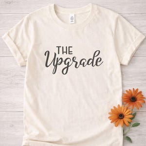 May include: Cream-colored t-shirt with the words "THE Upgrade" in black script. Two orange flowers with dark centers are placed on the right side of the shirt, against a light wood-grain background. The shirt has rolled-up sleeves.