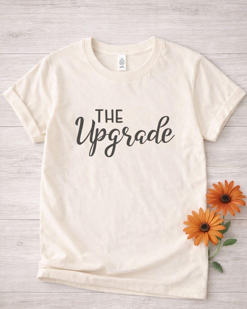 May include: Cream-colored t-shirt with the words "THE Upgrade" in black script. Two orange flowers with dark centers are placed on the right side of the shirt, against a light wood-grain background. The shirt has rolled-up sleeves.