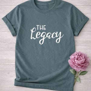 May include: A teal-colored t-shirt with rolled-up sleeves. The white cursive text "THE Legacy" is printed on the front. A pink peony flower with green leaves is placed on the right side of the shirt.