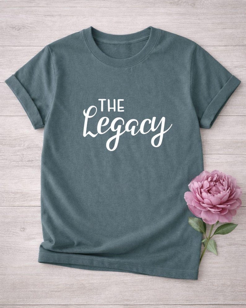 May include: A teal-colored t-shirt with rolled-up sleeves. The white cursive text "THE Legacy" is printed on the front. A pink peony flower with green leaves is placed on the right side of the shirt.
