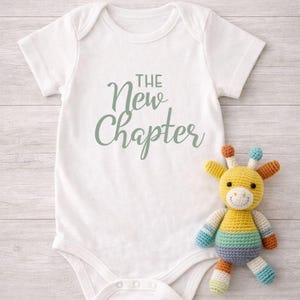 May include: A white baby onesie with the text "The New Chapter" in a soft green script. A colorful, crocheted giraffe toy with yellow, blue, orange, and green accents sits beside the onesie, creating a cheerful scene.