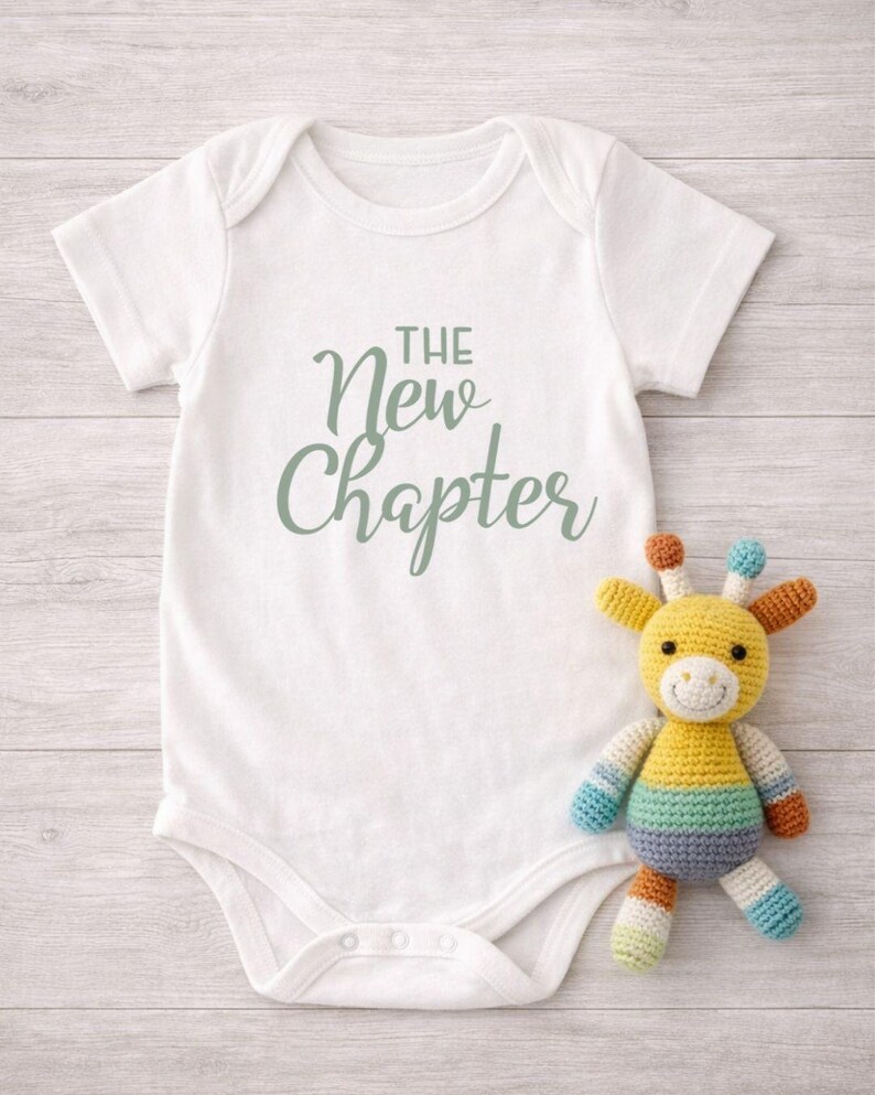 May include: A white baby onesie with the text "The New Chapter" in a soft green script. A colorful, crocheted giraffe toy with yellow, blue, orange, and green accents sits beside the onesie, creating a cheerful scene.