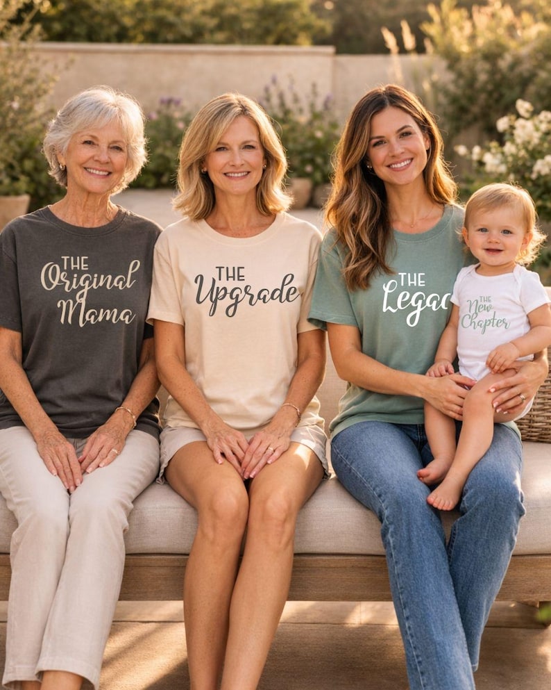 May include: Four people wearing matching shirts with text: "The Original Mama", "The Upgrade", "The Legacy", and "The New Chapter". The group is sitting on a bench outdoors. The shirts are various colors, including gray, beige, and green.