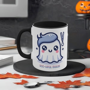 Pode incluir: Caneca de cerâmica branca com alça e interior pretos. A caneca apresenta um fantasma de desenho animado com cabelo azul e tesoura, com o texto "BOO-tiful Styles" em rosa e azul. A caneca está em um prato preto com folhas laranja.