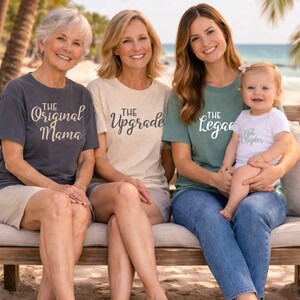 May include: Four people, including a baby, sit on a wooden bench near a beach. Each person wears a t-shirt with a different phrase: "The Original Mama," "The Upgrade," "The Legacy," and "The New Chapter."