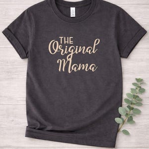May include: Dark gray t-shirt with rolled sleeves. The text "THE Original Mama" is printed in a light brown, cursive font across the front. The shirt is made of a soft material and is suitable for casual wear.