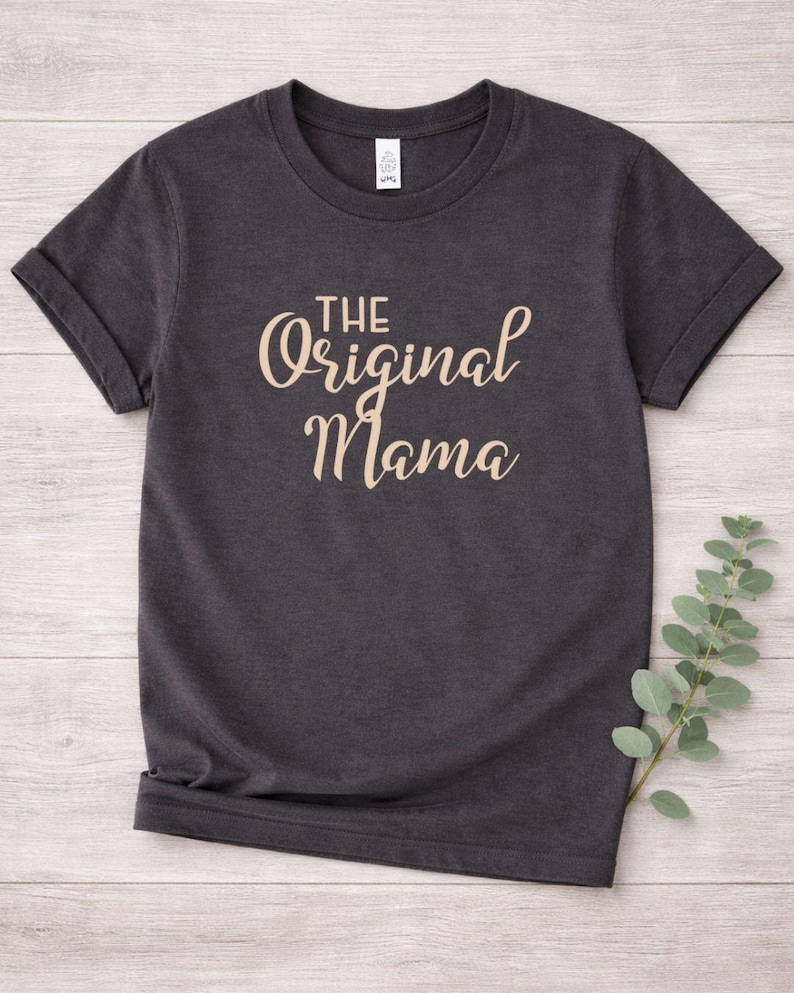 May include: Dark gray t-shirt with rolled sleeves. The text "THE Original Mama" is printed in a light brown, cursive font across the front. The shirt is made of a soft material and is suitable for casual wear.