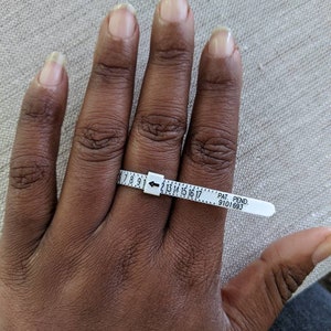 May include: A white plastic ring sizer with black markings from size 7 to 17. The sizer is on a person's finger. The text on the sizer reads "PAT. PEND. 9101693".