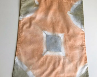 May include: A peach-colored tote bag with a geometric pattern in white, gray, and blue. The bag has matching peach-colored handles and is made of a soft, possibly cotton, fabric. The design is centered on the bag.