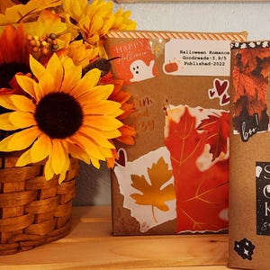 May include: A brown woven basket filled with yellow and orange sunflowers next to two books wrapped in brown paper. The books feature stickers of ghosts, red leaves, and text including "Halloween Romance," "Paranormal Horror," and "Spooky Season."