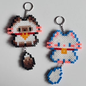 May include: Two handmade keychains. One is a brown and white owl with a red collar and a brown acorn charm. The other is a white and blue cat with a red collar and a blue heart charm. Both keychains are made of colorful beads.