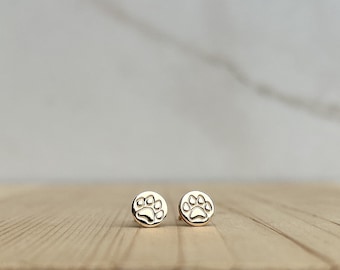May include: A pair of gold-colored stud earrings. Each earring features a circular design with a paw print in the center. The earrings are displayed on a light-colored wooden surface.