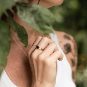 May include: A close-up of a person's hand wearing a gold ring with a black gemstone. The ring is on the index finger and is surrounded by greenery.