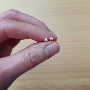 May include: A pair of silver triangle stud earrings. The earrings are small and delicate, and they are held in a person's hand.
