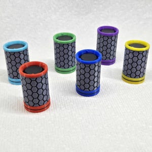 Könnte beinhalten: Sechs zylindrische Objekte mit einem grauen Wabenmuster und farbigen Rändern. Die Ränder sind in verschiedenen Farben: Rot, Blau, Grün, Lila, Gelb und Türkis. Die Oberseiten sind schwarz.