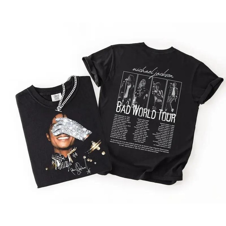 May include: Two black t-shirts featuring Michael Jackson. One shirt displays a portrait with a jeweled glove, the other has a "BAD WORLD TOUR" graphic with tour dates. A silver chain necklace is draped over the first shirt.