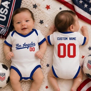 May include: A white baby bodysuit with blue trim, featuring the text Los Angeles and the number 00 in red on the front, and the text CUSTOM NAME and the number 00 in red on the back, displayed on two infants in a sports-themed setting.