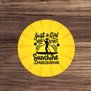 May include: A yellow circle with a black silhouette of a person paddleboarding. The text "Just a Girl Who Loves Sunshine & Paddleboarding" is written in black.
