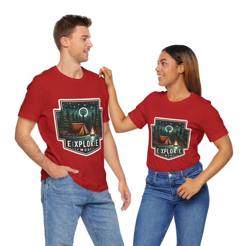 Puede incluir: Dos camisetas rojas con un dise&ntilde;o gr&aacute;fico de una escena de fogata bajo un cielo estrellado. El dise&ntilde;o incluye una tienda de campa&ntilde;a, &aacute;rboles y una fogata. El texto en las camisetas dice "Explore More".