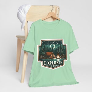 Puede incluir: Una camiseta verde claro con un gr&aacute;fico de un campamento bajo una luna creciente y estrellas. El gr&aacute;fico est&aacute; rodeado por un borde marr&oacute;n y el texto "EXPLORE MORE" est&aacute; debajo del gr&aacute;fico.