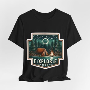 Puede incluir: Camiseta negra con un gr&aacute;fico de una escena de campamento con una tienda de campa&ntilde;a, una fogata y una luna creciente. El gr&aacute;fico est&aacute; rodeado por un borde verde y marr&oacute;n y el texto "EXPLORE MORE" est&aacute; debajo del gr&aacute;fico.