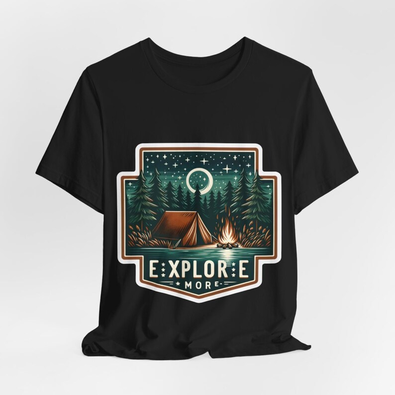 Puede incluir: Camiseta negra con un gr&aacute;fico de una escena de campamento con una tienda de campa&ntilde;a, una fogata y una luna creciente. El gr&aacute;fico est&aacute; rodeado por un borde verde y marr&oacute;n y el texto "EXPLORE MORE" est&aacute; debajo del gr&aacute;fico.