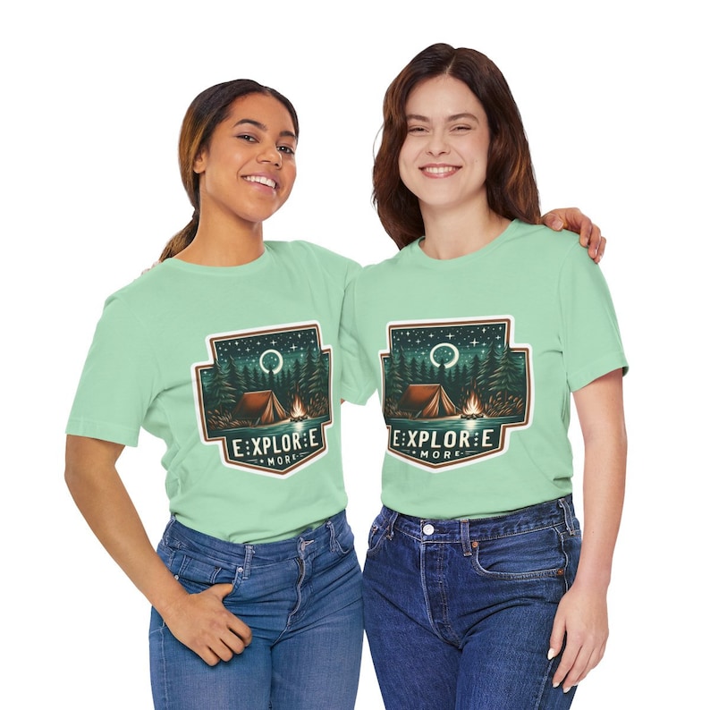 Puede incluir: Dos personas con camisetas verde claro con un gr&aacute;fico de escena de campamento y el texto "Explore More".