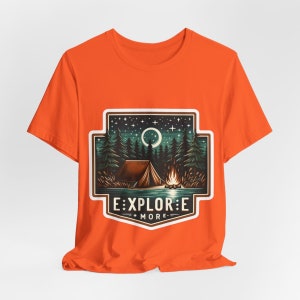 Puede incluir: Camiseta naranja con un gr&aacute;fico blanco de un campamento nocturno con una tienda de campa&ntilde;a, una fogata y una luna creciente. El gr&aacute;fico incluye el texto "EXPLORE MORE" en una pancarta blanca.