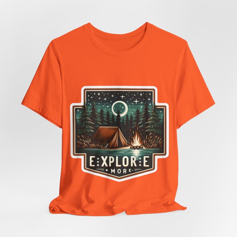 Puede incluir: Camiseta naranja con un gr&aacute;fico blanco de un campamento nocturno con una tienda de campa&ntilde;a, una fogata y una luna creciente. El gr&aacute;fico incluye el texto "EXPLORE MORE" en una pancarta blanca.