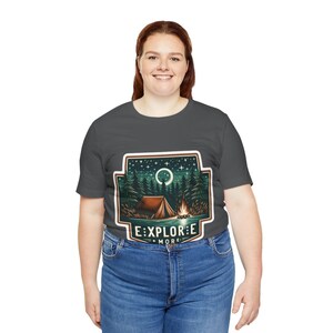 Puede incluir: Una camiseta gris oscuro con un gr&aacute;fico de un campamento nocturno con una tienda de campa&ntilde;a, una fogata y una luna creciente. El gr&aacute;fico est&aacute; rodeado por un borde blanco y el texto "EXPLORE MORE" est&aacute; debajo del gr&aacute;fico.