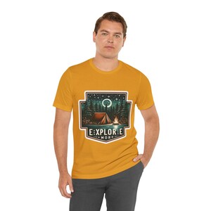Puede incluir: Una camiseta amarilla mostaza con un gr&aacute;fico de una escena de campamento. El gr&aacute;fico presenta una tienda de campa&ntilde;a, una fogata, &aacute;rboles y una luna creciente. El texto "EXPLORE MORE" est&aacute; debajo del gr&aacute;fico.