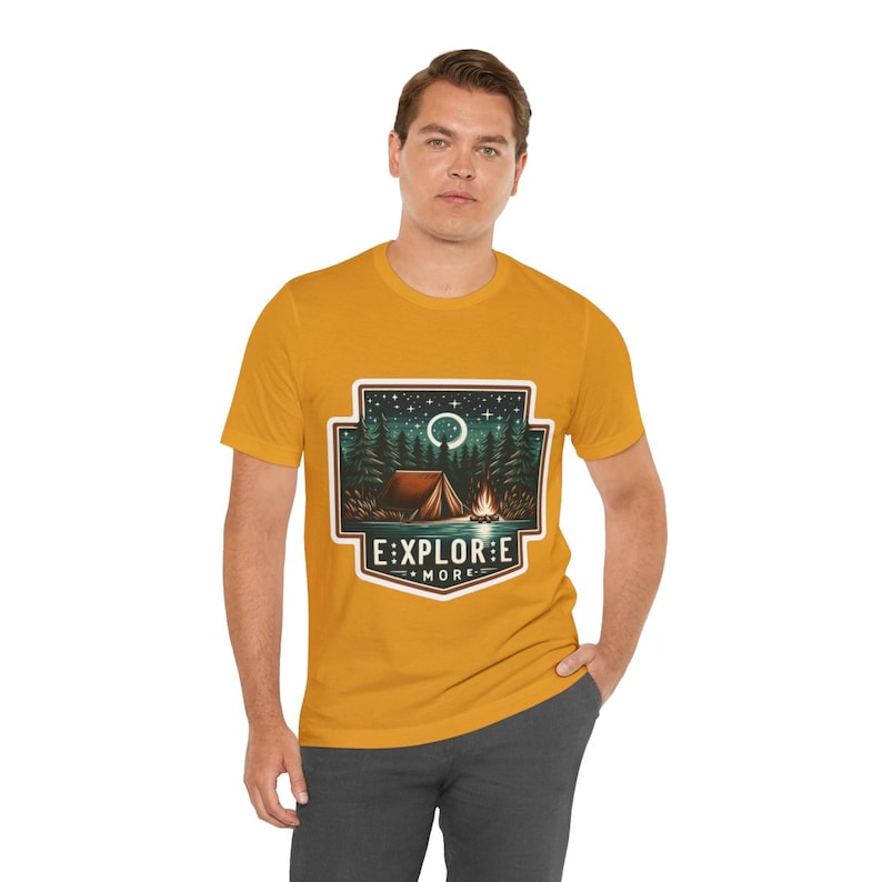 Puede incluir: Una camiseta amarilla mostaza con un gr&aacute;fico de una escena de campamento. El gr&aacute;fico presenta una tienda de campa&ntilde;a, una fogata, &aacute;rboles y una luna creciente. El texto "EXPLORE MORE" est&aacute; debajo del gr&aacute;fico.