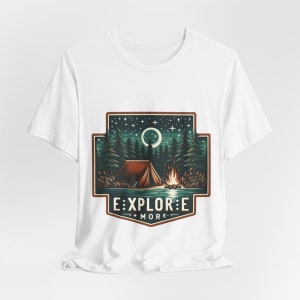 Puede incluir: Camiseta blanca con un dise&ntilde;o gr&aacute;fico de un campamento nocturno con una tienda de campa&ntilde;a, una fogata y una luna creciente. El gr&aacute;fico est&aacute; rodeado por un borde marr&oacute;n y el texto "EXPLORE MORE" est&aacute; debajo del gr&aacute;fico.