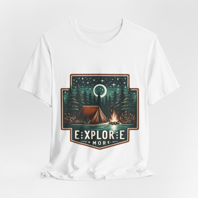 Puede incluir: Camiseta blanca con un dise&ntilde;o gr&aacute;fico de un campamento nocturno con una tienda de campa&ntilde;a, una fogata y una luna creciente. El gr&aacute;fico est&aacute; rodeado por un borde marr&oacute;n y el texto "EXPLORE MORE" est&aacute; debajo del gr&aacute;fico.