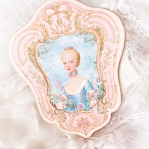 Könnte beinhalten: Ein rosa verzierter Handfächer mit einem Porträt einer Frau in einem blauen Kleid, eingebettet in einen dekorativen Rahmen. Der Fächer ist mit floralen und goldenen Akzenten verziert und mit einem weißen Band und einem dekorativen Anhänger gebunden.