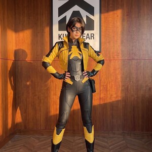 May include: A superhero costume featuring a black and yellow fitted bodysuit, a matching yellow and black cropped jacket, black gloves, and black boots. The outfit includes a black eye mask, a belt with a central emblem, and a small black side pouch.
