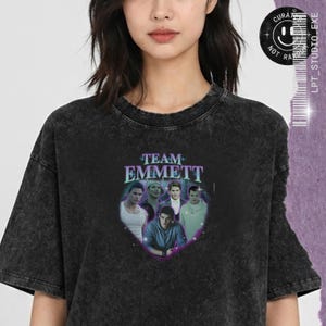 以下が含まれることがあります： 紫と青の「TEAM EMMETT」というテキストがプリントされた、黒いアシッドウォッシュ加工のTシャツ。デザインには、ハート型のフレームの中に5人の人物のコラージュが含まれており、小さな白い星と紫色の輝きのエフェクトがアクセントになっています。