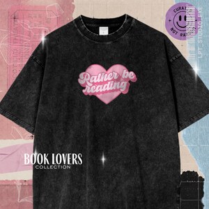 Peut inclure: Un t-shirt noir délavé à l'acide avec un motif de cœur rose sur la poitrine. À l'intérieur du cœur, le texte blanc indique "Rather be reading" dans une police cursive. Le t-shirt a un col rond et des manches courtes, avec une étiquette visible.