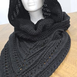 May include: A black hooded scarf, hand-knitted with a thick yarn. The hood is attached to a triangular scarf, with a textured pattern and openwork details. The scarf is displayed on a white mannequin head, set against a light background.